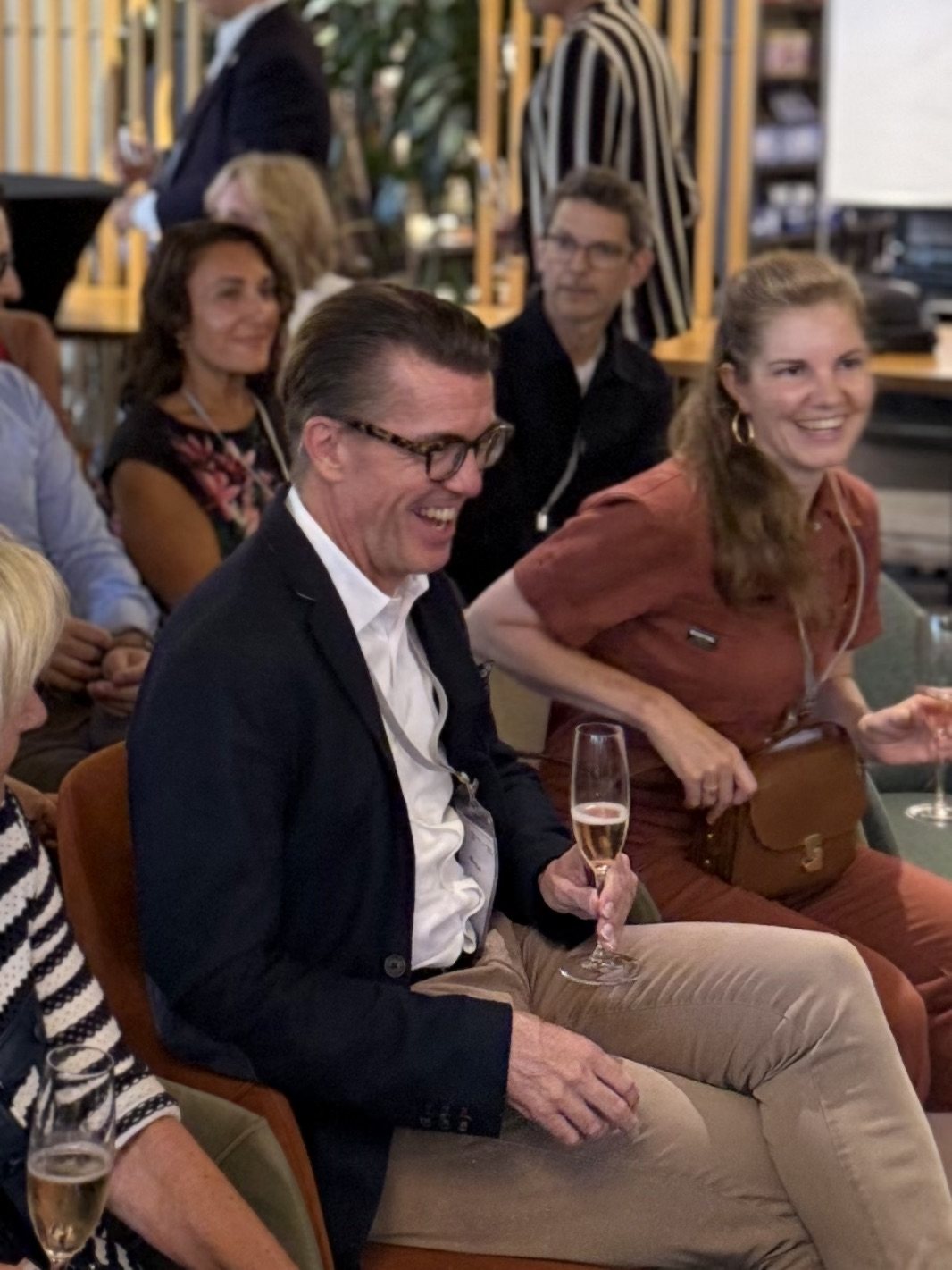 Gäste lachen bei einem Anlass, ein Mann mit Brille hält ein Glas